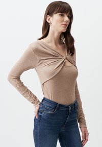 Top a maniche lunghe beige con design frontale attorcigliato e hardware dorato, abbinato a jeans skinny blu scuro. Texture liscia.