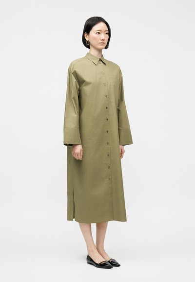 Γυναίκα που στέκεται φορώντας ένα μακρύ πουκαμισοφόρεμα σε ελιά πράσινο με κουμπιά, μακριά μανίκια και μαύρα μυτερά επίπεδα παπούτσια σε λευκό φόντο.
