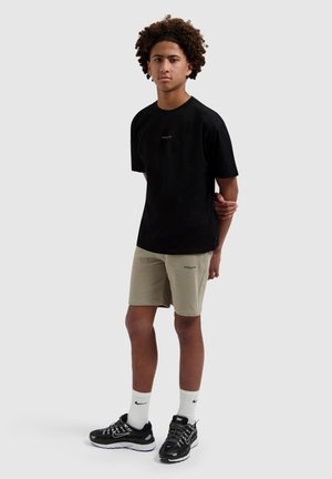 Jonge man met krullend haar staat, draagt een zwart T-shirt, beige korte broek, witte Nike-sokken en zwarte Nike-sneakers tegen een effen achtergrond.