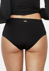 Femme portant un sous-vêtement noir taille haute et un haut noir à manches longues, montrant le bas du dos et le haut des jambes sur fond blanc.