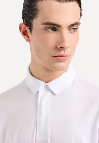 Polo shirt blanc avec un col classique, comportant trois boutons et un logo subtil sur la poitrine. Tissu lisse et design ajusté.