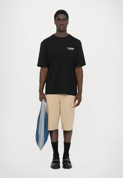 T-shirt noir oversize avec du texte blanc, shorts beiges avec poches, tenant un sac en denim, chaussettes noires et mocassins brillants.