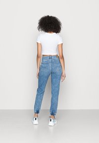 Žena s kudrnatými vlasy stojí s obratem, má na sobě bílý zkrácený tričko, modré džíny s vysokým pasem a elastickými manžetami a bílé tenisky.