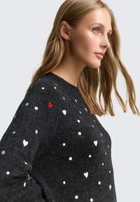 Schwarzer Pullover mit weißen Punkten und Herzmustern. Verfügt über einen kleinen roten Herzakzent an der Schulter. Weicher Stoff, lockere Passform.