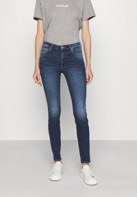 Jeans ajustados de denim azul con una cintura alta y un acabado desgastado. Combinados con una camiseta gris y zapatillas blancas, de pie sobre un fondo neutral.