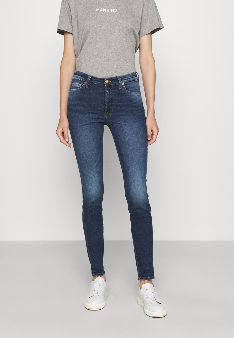 Jeans ajustados de denim azul con una cintura alta y un acabado desgastado. Combinados con una camiseta gris y zapatillas blancas, de pie sobre un fondo neutral.