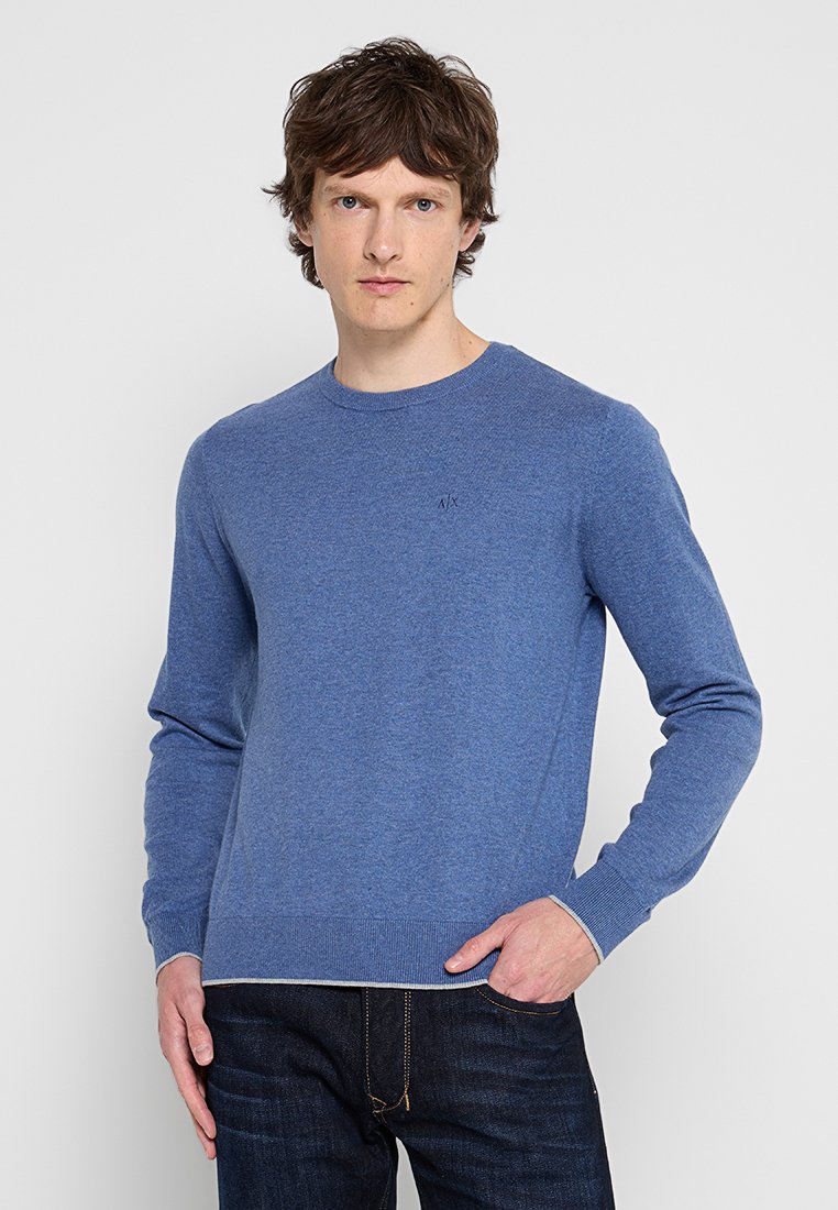 Blauer Rundhals-Pullover aus weichem Material. Hat lange Ärmel, gerippte Bündchen und dezente Markenkennung auf der Brust. Getragen mit dunklen Jeans.
