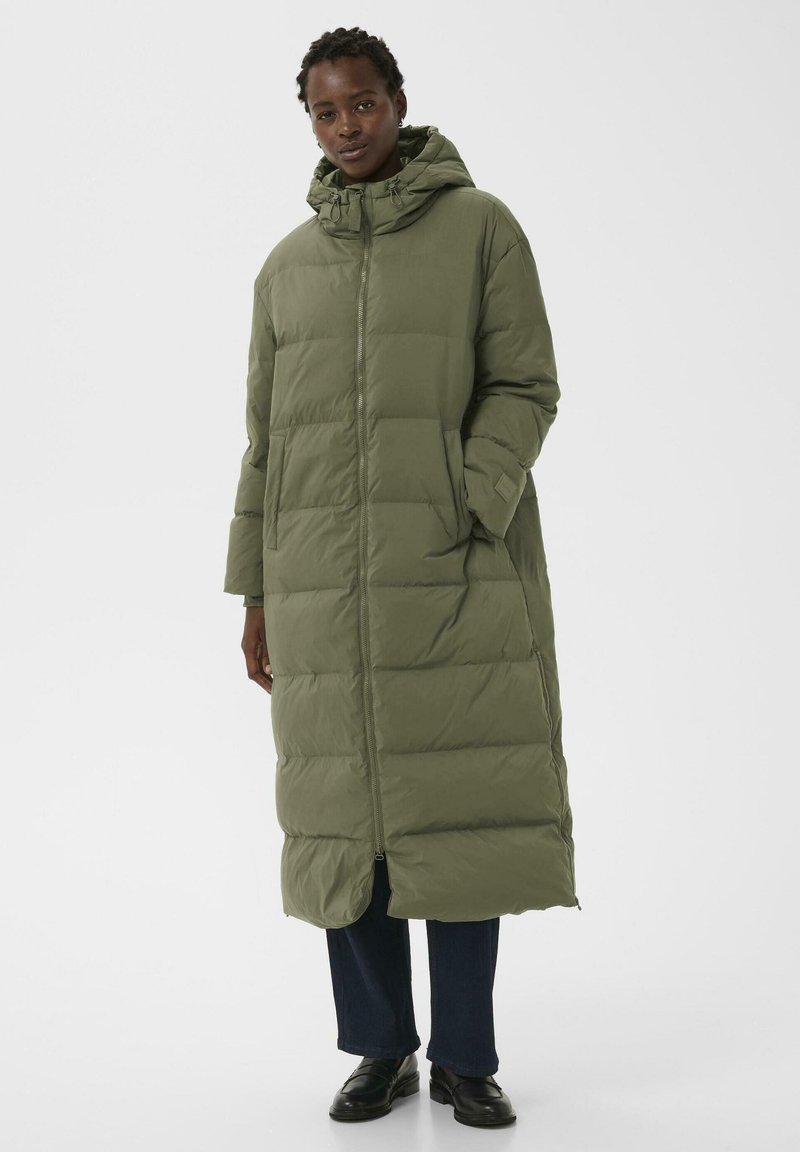 Donna che indossa un lungo piumino verde oliva con cappuccio, cerniera frontale e tasche laterali, abbinato a pantaloni scuri e scarpe nere.