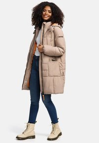 Beige pufferjack met een oranje binnenvoering, ritssluiting en capuchon. Voorzien van een voorzak en geribde manchetten. G gedragen met blauwe jeans en beige laarzen.