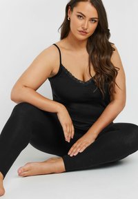 Svart linne med spetskant och tunna band tillsammans med svarta leggings. Mjukt tyg, figurnära design, modell sitter med bara fötter.