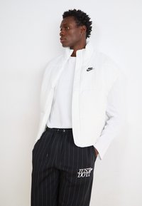 Λευκό γιλέκο πούπουλο με ψηλό γιακά και λογότυπο Nike, σε στρώσεις πάνω από ένα λευκό μπλουζάκι μακρυμάνικο. Συνδυασμένο με μαύρο παντελόνι με ρίγες και λεπτομέρεια λογότυπου.