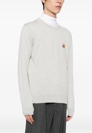Persona che indossa un maglione grigio chiaro con un piccolo logo arancione di volpe sul petto, un dolcevita bianco sotto e pantaloni grigio scuro.