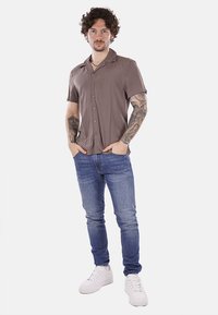 Camicia a maniche corte in tessuto marrone chiaro con colletto, chiusura con bottoni, abbinata a jeans blu e sneakers bianche.