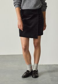 Black corduroy wrap skirt with a smooth texture, knee-length, featuring a front flap panel and paired with black loafers and gray socks.