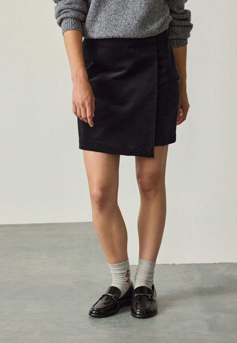 Black corduroy wrap skirt with a smooth texture, knee-length, featuring a front flap panel and paired with black loafers and gray socks.