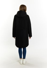 Sort lang frakke med hætte, glat tekstur, dobbeltlagd bund og sidelommer, parret med blå skinny jeans og hvide sneakers.