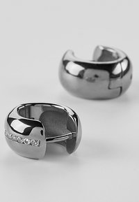 Orecchini a cerchio in metallo argento con una forma arrotondata e una fila di piccoli cristalli trasparenti su un lato.