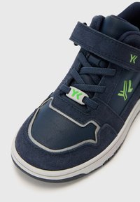 Navy blue sneaker with a suede and synthetic upper, lace and Velcro strap closure, and green accents on logo and design details.