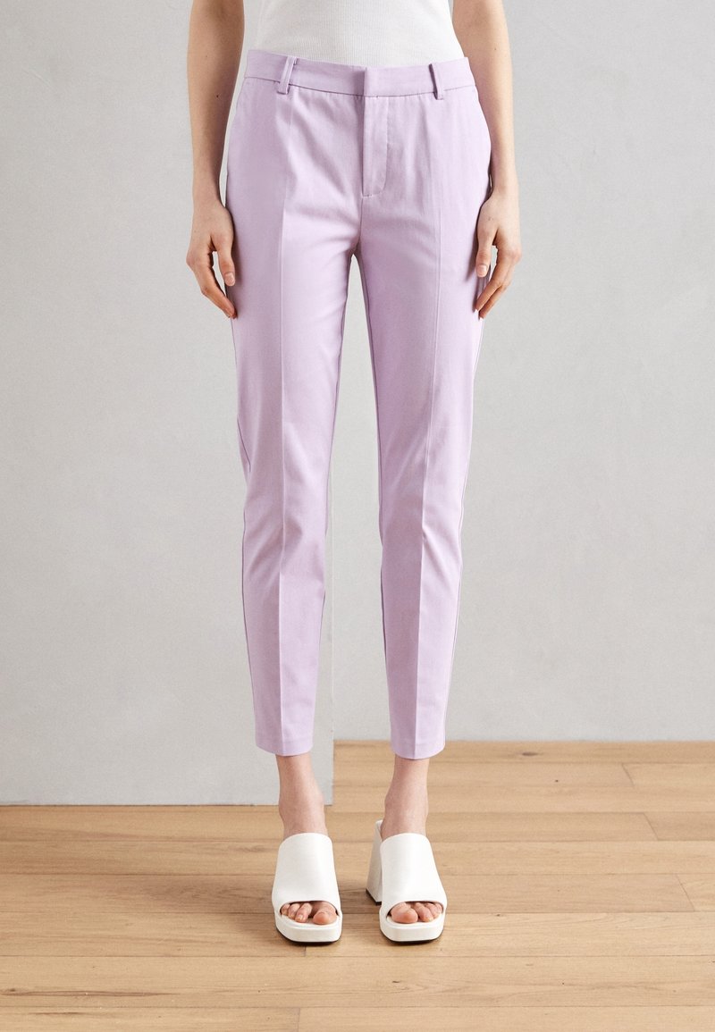 Personne portant un pantalon lilas ajusté longueur cheville et des sandales blanches à talon bloc ouvertes devant, debout sur un sol en bois contre un mur gris.