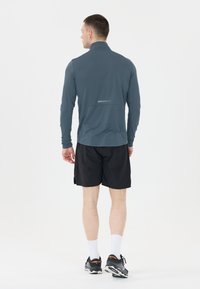 Långärmad sporttröja i grått med hög krage, utrustad med en reflekterande detalj på baksidan. Bärs med svarta shorts och löparskor.