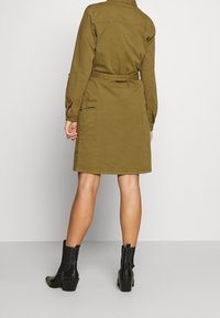 Vestito camicia verde oliva con cintura, maniche lunghe, tasche laterali e taglio dritto. Abbinato a stivali neri alla caviglia con tacco a blocco.