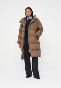 Př padded hnědý dlouhý kabát s vysokým límcem, zapínáním na patentky a bočními kapsami, s hladkým, texturovaným povrchem.
