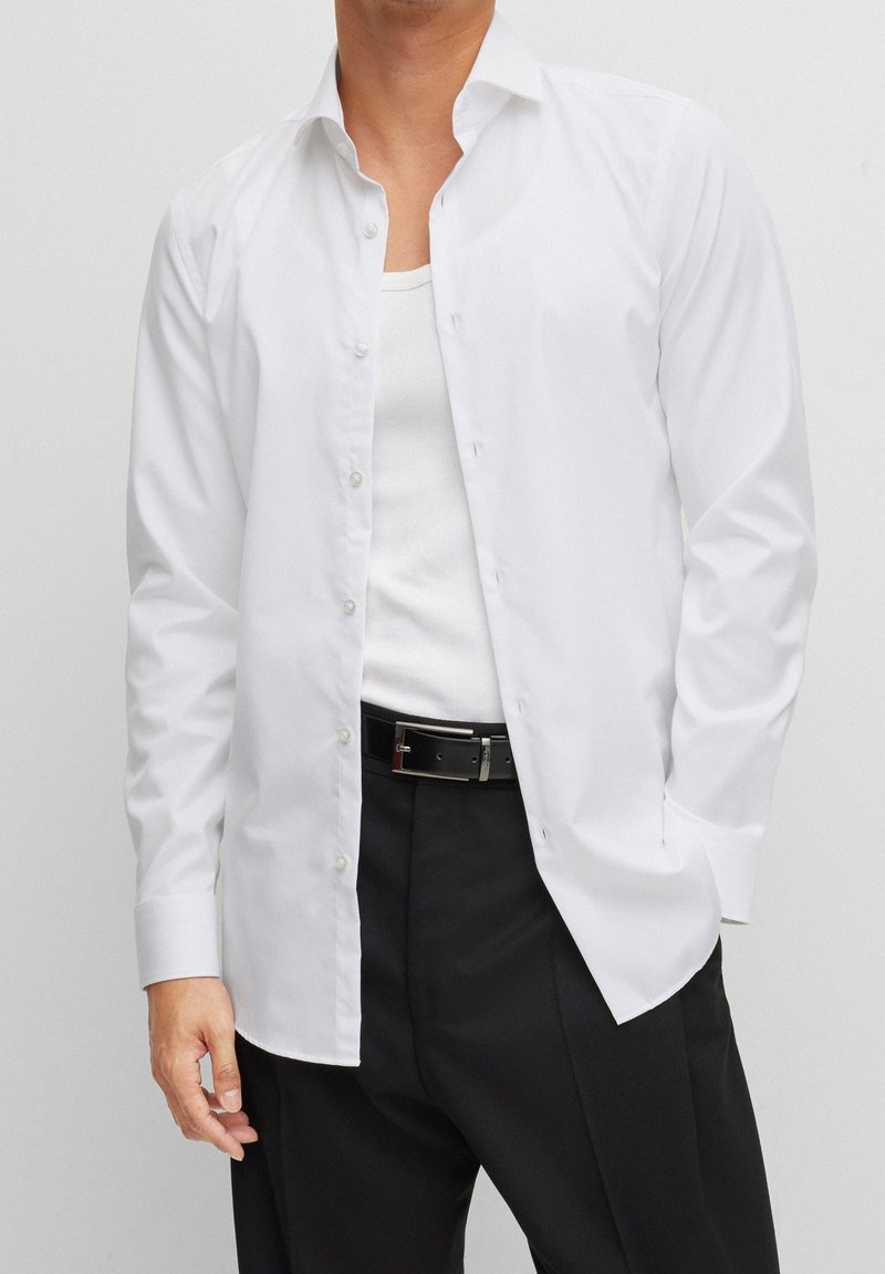 Homme portant une chemise blanche déboutonnée par-dessus un débardeur blanc, pantalon noir et ceinture noire, debout avec une main dans la poche.