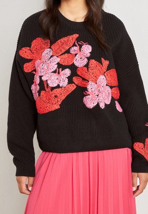 Zwarte gebreide trui met levendige rode en roze bloemenborduursels. Heeft een ontspannen pasvorm en geribde manchetten. Gecombineerd met een geplooide roze rok.