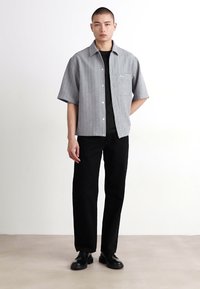 Cămașă gri cu mânecă scurtă și nasturi, cu dungi verticale, purtată peste un tricou negru, asortată cu pantaloni negri drepti și pantofi negri.