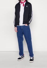 Svart zip-up jacka med vita ränder, vit t-shirt, blå jeans och svarta sneakers med vita sulor. Röd bandana runt halsen.