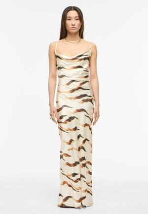 Žena stojaca v krémových šatách bez rukávov s abstraktnými hnedými a čiernymi pruhmi tigra, dĺžka až po zem.
