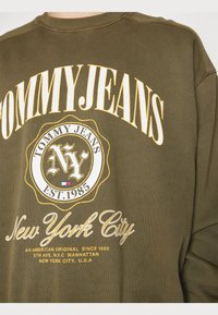 Felpa verde oliva con un notevole design del logo giallo e bianco, con il testo "TOMMY JEANS" e "New York City". Tessuto morbido e testurizzato.