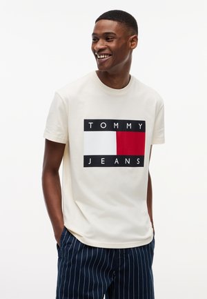 Homme souriant portant un t-shirt blanc Tommy Jeans avec un logo rouge, blanc et noir, et un pantalon bleu marine à fines rayures, les mains dans les poches.