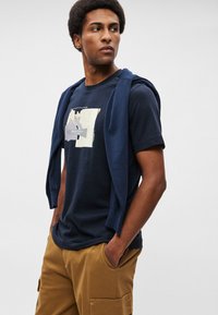 T-shirt azul marinho com estampado gráfico, acompanhado de um suéter azul marinho pousado sobre os ombros. Usado com calças cargo cáqui, com bolsos.