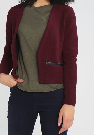 Personne portant une veste texturée bordeaux avec une poche bordée de noir, une chemise vert olive et un pantalon sombre, une main dans la poche.