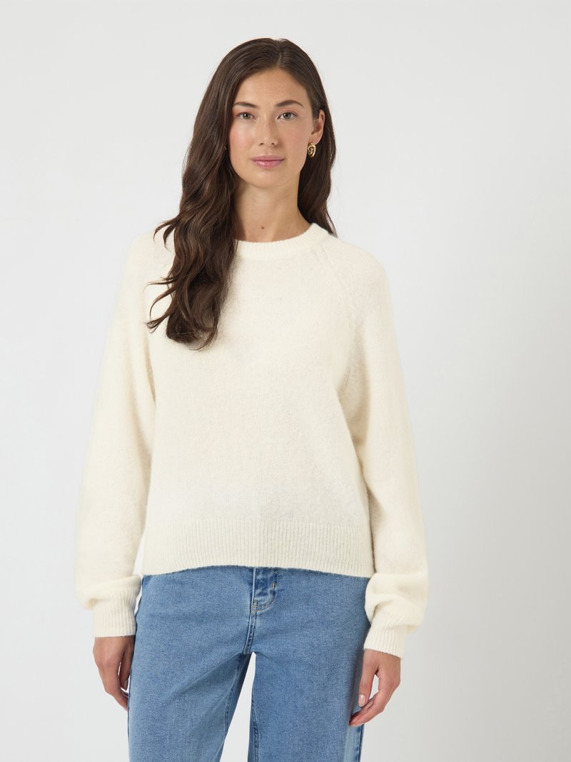 Kobieta z długimi brązowymi włosami, nosząca złote kolczyki w kształcie kół, biały sweter z dzianiny i niebieskie dżinsy, stojąca na jednolitym jasnym tle.