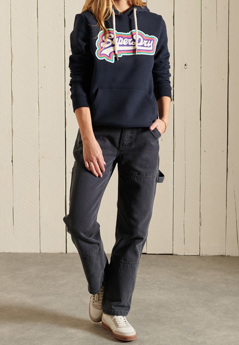 Femme portant un sweat à capuche bleu marine Superdry, un pantalon cargo noir, des baskets blanches, debout devant un fond en panneaux de bois clair.