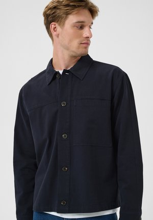 Joven con una chaqueta azul marino oscuro abotonada y con cuello, mirando hacia un lado, sobre un fondo claro y liso.