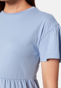 Vaaleansininen lyhythihainen paita, joka on valmistettu pehmeästä puuvillasta. Siinä on pyöreä kaula-aukko ja poimutetut hihat. Sujuva pinta, jossa on hienovaraisia ompeluyksityiskohtia.