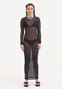 Schwarzes, durchsichtiges Langarmkleid mit floralen Mustern, hohem Kragen und seitlichen Schlitzen. Wird über einem schwarzen Dessous-Set getragen und mit schwarzen Sandalen kombiniert.