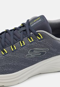 Scarpa sportiva blu navy con tomaia in rete, dettagli testurizzati, accenti gialli e suola in schiuma bianca. Presenta un logo prominente sul lato e nei lacci.