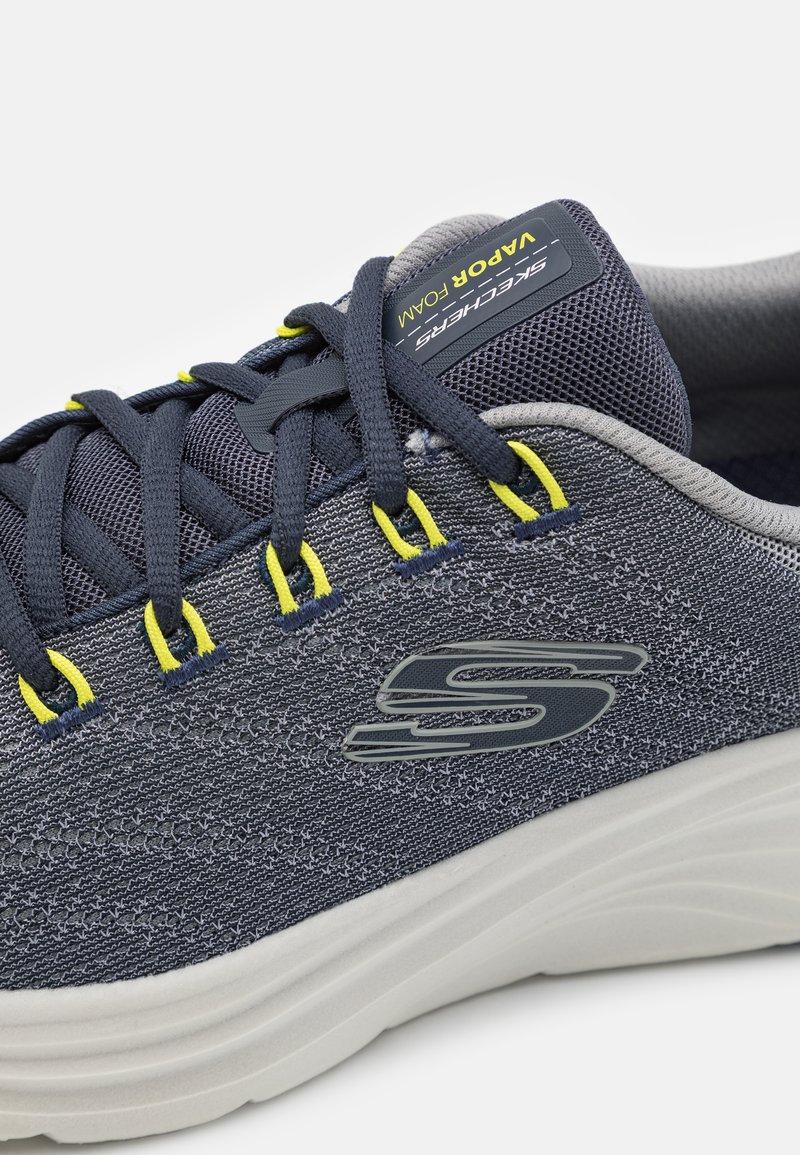 Granatowe buty sportowe z siatkowym cholewką, teksturowanymi detalami, żółtymi akcentami oraz białą piankową podeszwą. Zawierają wyraźne logo z boku i na sznurowadłach.