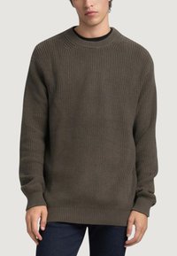 Suéter de punto texturizado en verde oscuro con cuello redondo y puños y bajo acanalados, combinado con jeans azul oscuro.