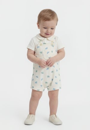 Νήπιο με ανοιχτό καφέ μαλλιά, φορώντας λευκό πουκάμισο με γιακά και κρεμ ολόσωμη φόρμα με εκτυπώσεις δεινοσαύρων σε μπλε-πράσινο, όρθιο και με τα χέρια κλεισμένα.