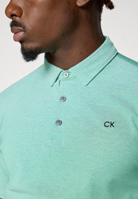 Grünes Polo-Shirt aus strukturiertem Stoff, mit kurzen Ärmeln, einem Kragen und drei grauen Knöpfen. Kleines schwarzes "CK"-Logo auf der Brust.