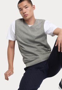 Gilet grigio a maglia con scollo a V, indossato sopra una maglietta bianca. Il modello indossa pantaloni a righe blu navy. Tessuto liscio, vestibilità standard.