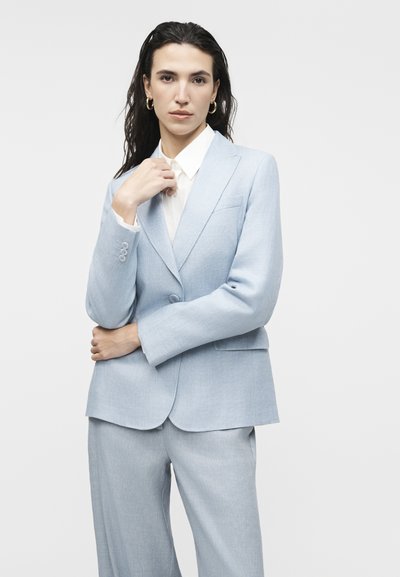 Γυναίκα με σκούρα μαλλιά που φορά χρυσούς κρίκους στα αυτιά και ανοιχτό μπλε κοστούμι με λευκό πουκάμισο, στέκεται με το ένα χέρι στον γιακά της.