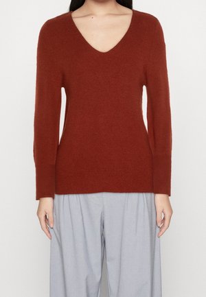 Personne portant un pull à manches longues couleur rouille avec col en V et un pantalon ample gris clair, debout devant un fond uni.
