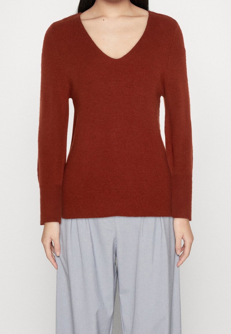 Personne portant un pull à manches longues couleur rouille avec col en V et un pantalon ample gris clair, debout devant un fond uni.