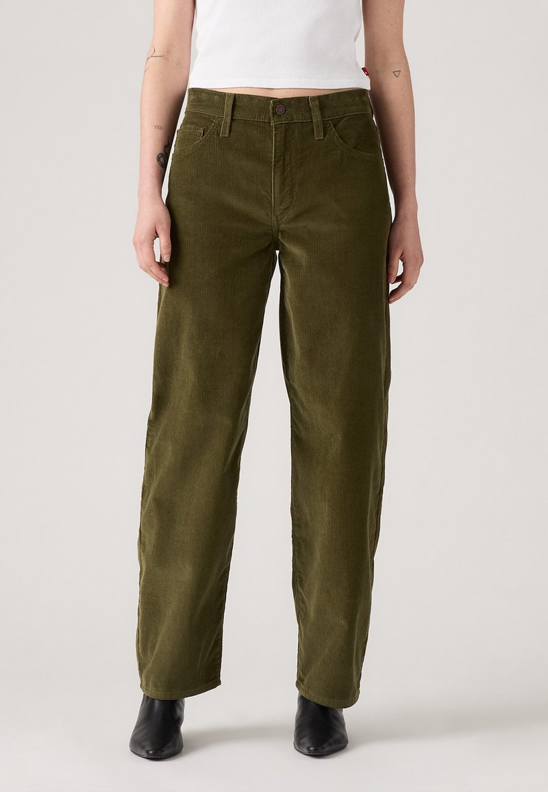 Personne portant un pantalon en velours côtelé vert olive à jambes droites, un top court blanc et des bottines noires, debout devant un fond uni.